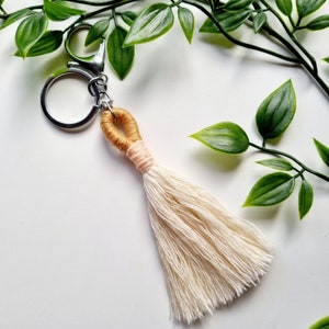 May include: A white tassel keychain with a gold and pink accent. The keychain has a silver metal ring.