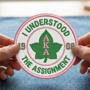 May include: Embroidered patch with a pink and green design. The patch features the text "I UNDERSTOOD THE ASSIGNMENT" in green, surrounding a green leaf with the letters "AKA" in pink. The numbers "19" and "08" are also included.