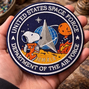 May include: Circular navy blue patch with white border featuring Snoopy in an orange spacesuit, a silver star, and the text "UNITED STATES SPACE FORCE DEPARTMENT OF THE AIR FORCE". Includes a globe, a silver star, and an orange planet.