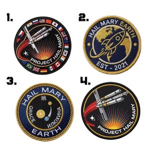 May include: Four circular embroidered patches with space-themed designs. One patch features a rocket and the text "HAIL MARY EARTH EST - 2021". Other patches include rockets, flags, and the words "PROJECT HAIL MARY".
