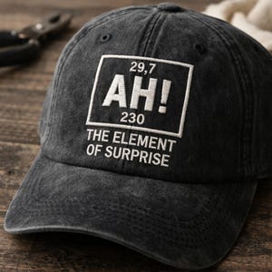 May include: A dark gray baseball cap with a white embroidered design. The design includes the text "AH!" with numbers and the phrase "THE ELEMENT OF SURPRISE". The cap is made of a worn, textured fabric.