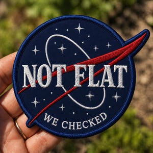 May include: A navy blue embroidered patch with the words "NOT FLAT" in white, and "WE CHECKED" in white. The patch features a red swoosh and white stars, resembling a NASA-style design.