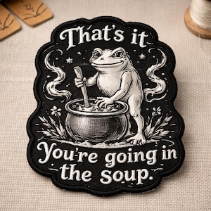 May include: Black and white embroidered patch featuring a frog stirring a cauldron. The text "That's it, You're going in the Soup" is embroidered in white. The patch has a decorative border.