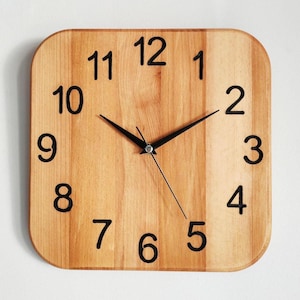 May include: Square wooden wall clock with rounded corners. The clock face is light brown with black numerals and black hands. The clock shows the time as 9:53. A simple, modern design for home decor.
