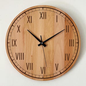May include: A round wooden wall clock with a light brown finish. The clock face features black Roman numerals and black clock hands. The outer edge has a decorative black border. The clock is a decorative home decor item.
