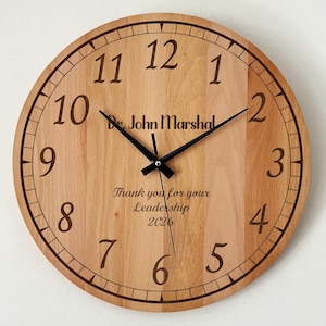 May include: A round wooden clock with black hands and numerals. The clock face has the text "Dr. John Marshal" and "Thank you for your Leadership 2026". The clock is made of light-colored wood.