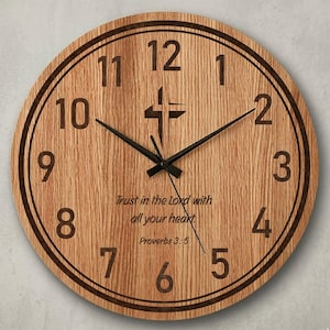 May include: Round wooden wall clock with a light brown finish. The clock features black numerals, hands, and a cross symbol at the 12 o'clock position. The text "Trust in the Lord with all your heart Proverbs 3:5" is inscribed.