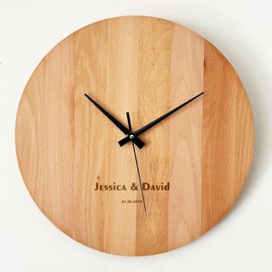 May include: A round wooden clock with black hands and the names "Jessica & David" engraved on the face, along with the date "01.06.2019". The clock has a light brown wood grain.