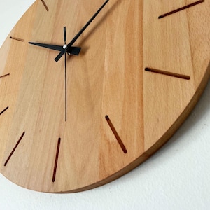 May include: A wooden wall clock with a light brown finish. The clock has black hands and simple, cut-out hour markers. The clock's design is minimalist and modern, suitable for any room. The clock's diameter is approximately 12 inches.