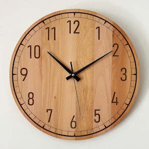 May include: A round wooden wall clock with a light brown face. The clock features black hands and brown numerals. The outer edge has a decorative black border. The clock is a simple, modern design.