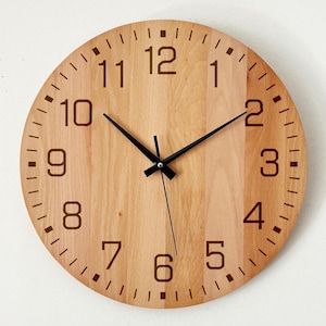 May include: A round wooden wall clock with a light brown finish. The clock features black hands and large, dark brown numerals. The clock's face has small, dark brown markings for each minute.
