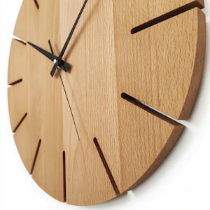 May include: A round wall clock made of light-colored wood with a visible grain pattern. The clock face features thin, dark-colored metal hands and deep, narrow slots cut into the wood at the hour positions instead of traditional numbers.
