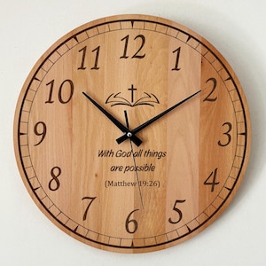 Könnte beinhalten: Runde Wanduhr aus hellem Holz mit schwarzen Zeigern und Ziffern. Das Zifferblatt zeigt den Text "With God all things are possible (Matthew 19:26)" und ein Kreuz über einem offenen Buch.