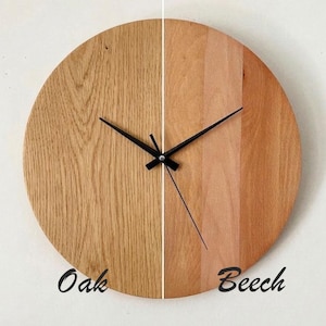 May include: A round wooden wall clock with a split design, featuring oak on the left and beech on the right. The clock has black hands and the words "Oak" and "Beech" are written on the clock face.