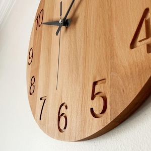 May include: A wooden wall clock with a light brown finish. The clock face features cut-out numerals and black clock hands. The numbers 5, 6, 7, 8, 9, and 10 are visible. The clock is mounted on a white wall.