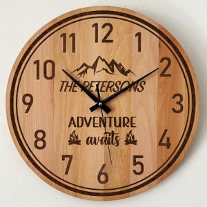 May include: A round wooden wall clock with a light brown grain finish, featuring black numerals from 1 to 12. The center displays a mountain graphic, the text THE PETERSONS, the words ADVENTURE awaits, and two small campfire icons with black hands.