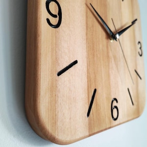 May include: A square wooden clock with a light brown finish. The clock face features black numerals at 3, 6, and 9 o'clock, with black lines marking the other hours. Black clock hands indicate the time.