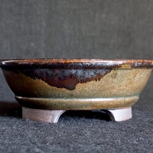 May include: A small, handcrafted ceramic bowl with a low profile and three small feet. The bowl has a mottled, earthy colour palette with shades of brown, green, and tan. The rim has a darker, glossy finish.