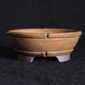 May include: A small, round, ceramic bonsai pot in light brown and green. The pot has a low profile with a slightly flared rim and three small, grey feet. The pot is designed for growing small plants, ideal for a miniature garden.