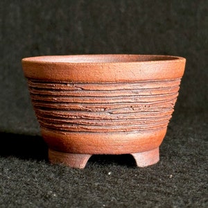 May include: A small, brown, ceramic bonsai pot with a textured, ribbed design and three short legs.