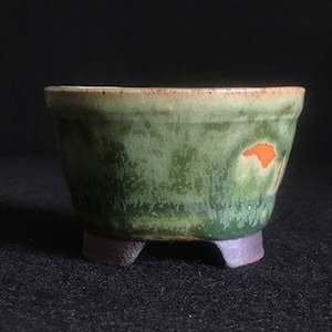 May include: A green ceramic bonsai pot with a brown rim and three small feet. The pot has a mottled green glaze with a small orange patch.