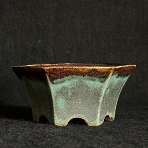 May include: A small, square bonsai pot with a unique design. The pot features a dark brown rim, a turquoise-green midsection, and a gray base. The pot has three small feet and a slightly flared shape. The pot is made of ceramic.