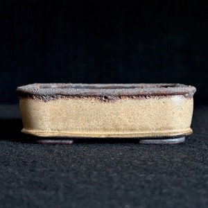 May include: A rectangular ceramic bonsai pot with a light brown glaze and a dark brown rim. The pot has three small feet on the bottom.