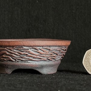 May include: A small, brown ceramic bonsai pot with a textured, woven design. The pot has three legs and is about the size of a British pound coin.