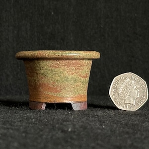 May include: A small, handcrafted ceramic bonsai pot with a flared shape and a rim. The pot features earthy tones of brown, green, and tan. A coin is next to the pot for size reference.