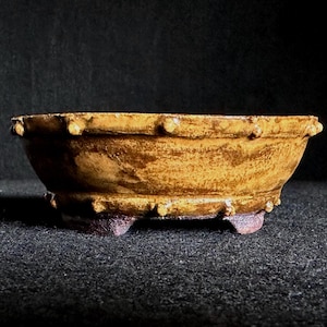 May include: A small, handcrafted ceramic bowl with a textured, earthy appearance. The bowl is primarily a warm, golden-brown colour with darker brown accents and small, rounded protrusions along the rim. It rests on three small feet.
