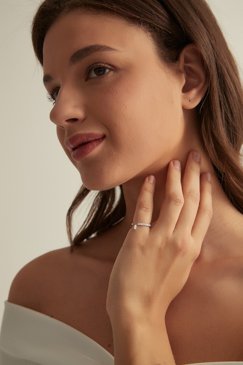 May include: A silver ring with a small diamond teardrop pendant. The ring is set with small diamonds and is worn on the ring finger of a person's hand.