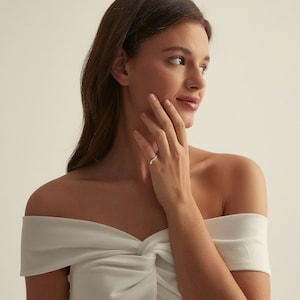 May include: A woman wearing a white off-the-shoulder top with a knotted detail, showing off a silver ring on her left ring finger.