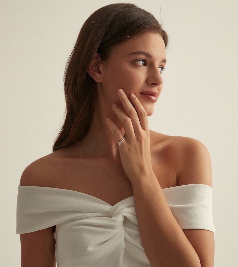 May include: A woman wearing a white off-the-shoulder top with a knotted detail, showing off a silver ring on her left ring finger.