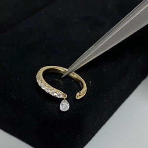 May include: A gold ring with a single dangling pear-shaped diamond. The ring is set with small diamonds around the band.