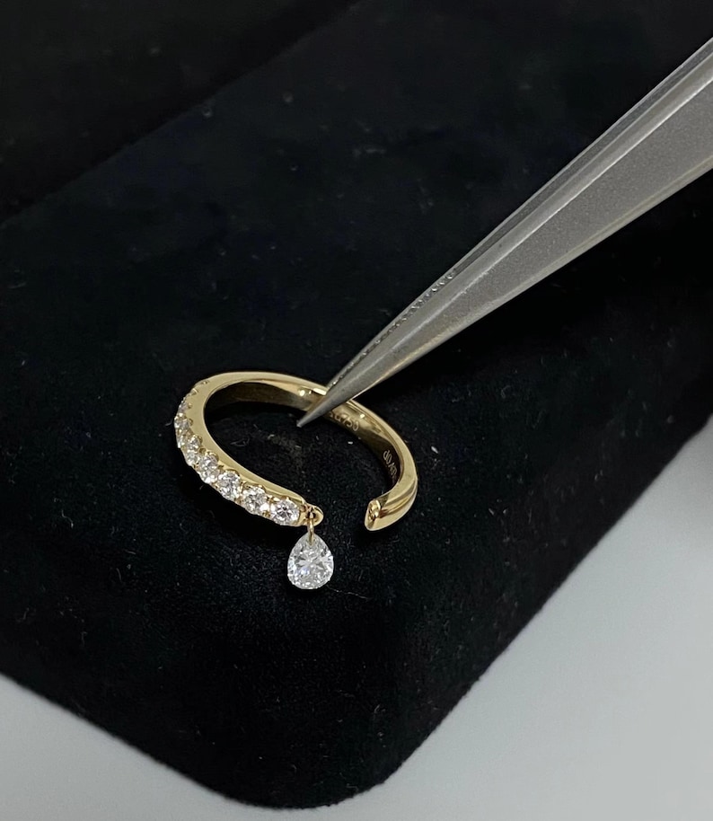 May include: A gold ring with a single dangling pear-shaped diamond. The ring is set with small diamonds around the band.