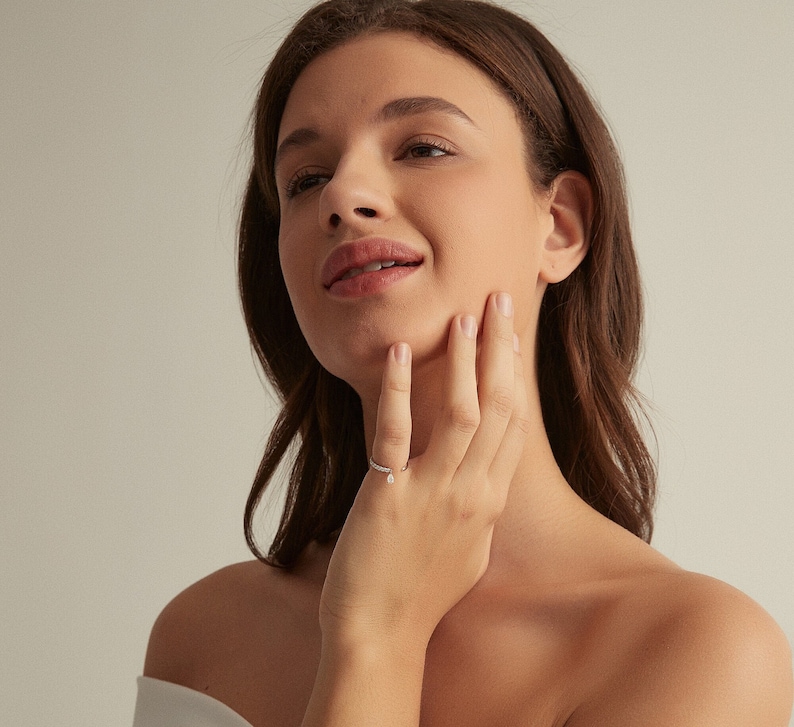 May include: A woman wearing a white off-the-shoulder top with her hand on her neck, showing off a delicate silver ring with a small diamond on her finger.