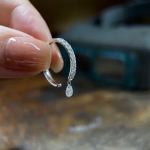 May include: A delicate silver ring with a single pear-shaped diamond dangling from the band. The ring is set with a row of smaller diamonds around the band.
