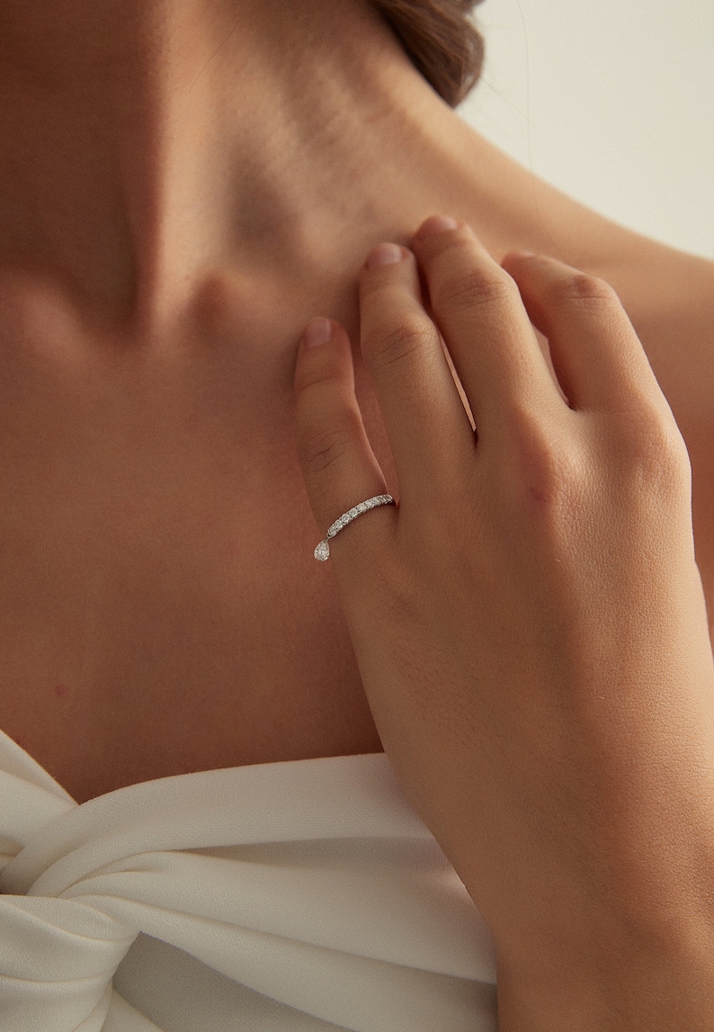 May include: A delicate silver ring with a single teardrop-shaped diamond dangling from the band. The ring is set with small diamonds that encircle the band.