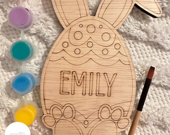 May include: Wooden bunny-shaped Easter egg craft with the name "EMILY" written on it. The craft is unfinished and ready to be painted with the included paint pots.