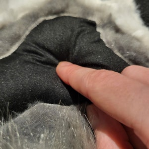 May include: Close-up of a grey and white furry paw with a black fabric pad. The pad is textured and has a slight indentation in the centre.