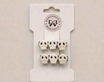 May include: Six white skull hair clips on a white card with the text "Dominique's Bowtique" in a circular logo. The clips are arranged in two rows of three, each skull detailed with eye sockets and a nose cavity.