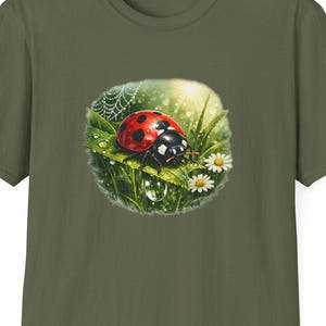Pode incluir: Uma t-shirt de manga curta verde-azeitona com uma impressão gráfica central de uma joaninha vermelha com pintas pretas pousada numa folha verde. O design inclui margaridas brancas, uma teia de aranha e uma gota de água sob a folha.