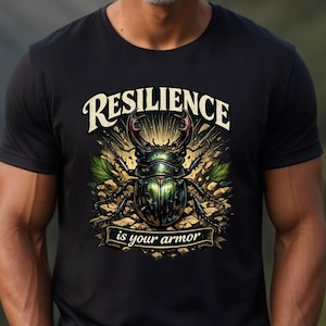 May include: Black t-shirt featuring a detailed illustration of a beetle with the words "RESILIENCE is your armor" in a decorative font. The beetle is depicted in shades of green, gold, and brown.