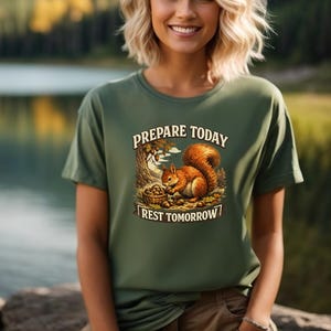 Op de afbeelding: Olijfgroen T-shirt met de tekst "Prepare Today, Rest Tomorrow" en een illustratie van een eekhoorn. De eekhoorn verzamelt noten. Het T-shirt wordt door een persoon gedragen.