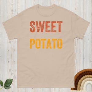 Può includere: T-shirt beige con la scritta "SWEET POTATO" in lettere maiuscole arancioni e gialle. Il testo ha un aspetto invecchiato. La maglietta ha un girocollo e maniche corte.