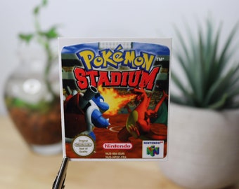 Op de afbeelding: Een rechthoekige sticker met de cover art van het videospel Pokemon Stadium. De afbeelding toont Blastoise en Charizard in een stadion met de tekst Pokemon Stadium, Nintendo en het Nintendo 64-logo duidelijk zichtbaar.