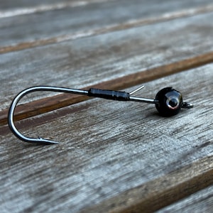 May include: A close-up of a fishing jig with a black, round head and a silver hook. The hook is curved with a sharp point. The jig is on a weathered wooden surface.