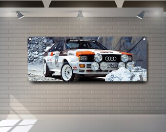Op de afbeelding: Een witte Audi Quattro rallyauto met oranje en zwarte accenten. De auto heeft een wit en zwart kenteken met de tekst "NM 61". De auto rijdt op een grindweg met rotsen en sneeuw op de achtergrond.