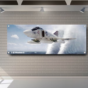 Op de afbeelding: Een witte F-4F Phantom II straaljager vliegt laag over de oceaan en creëert een grote waterstraal. Het vliegtuig heeft een donkerblauwe staart en het nummer "06" aan de zijkant.