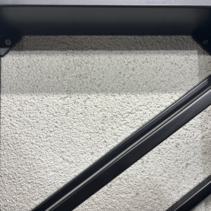 May include: Close-up of a black metal wine rack with a textured white background. The rack is made of two angled metal bars.